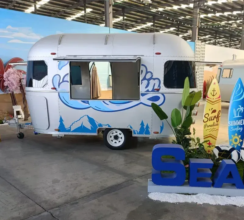 Weißer Imbisswagen mit blauer Ozean- und Berggrafik, offenem Bedienungsfenster, Surfbrettern.