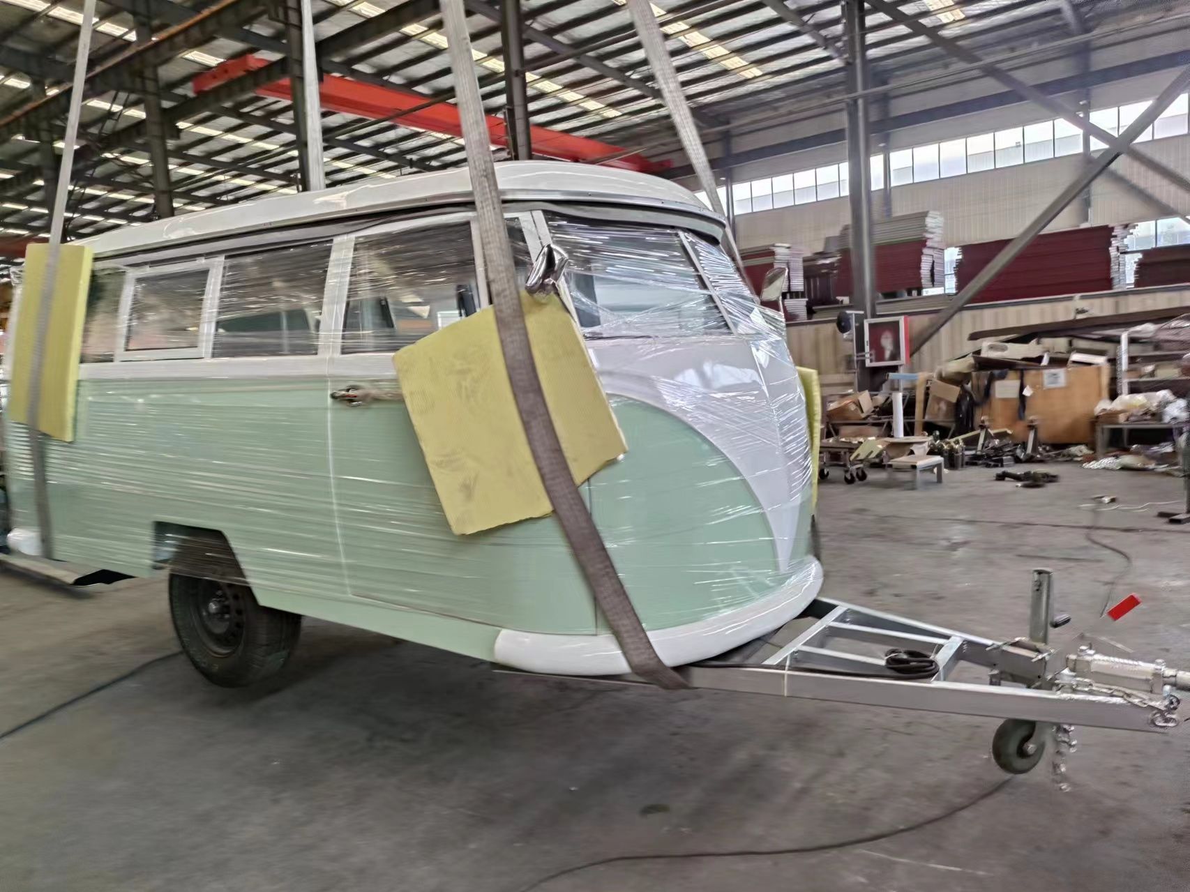 Ein mintgrüner Wohnwagenanhänger, der einem alten VW-Bus ähnelt, wird in einer Lagerhalle mit Gurten hochgehoben.