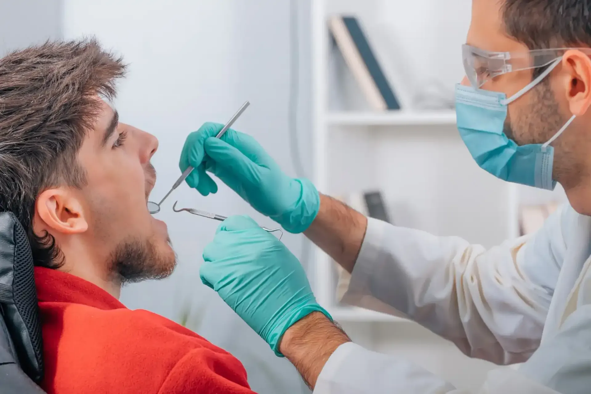 Dentista examinando la boca de un paciente con instrumental, usando guantes, mascarilla y gafas en un consultorio dental.