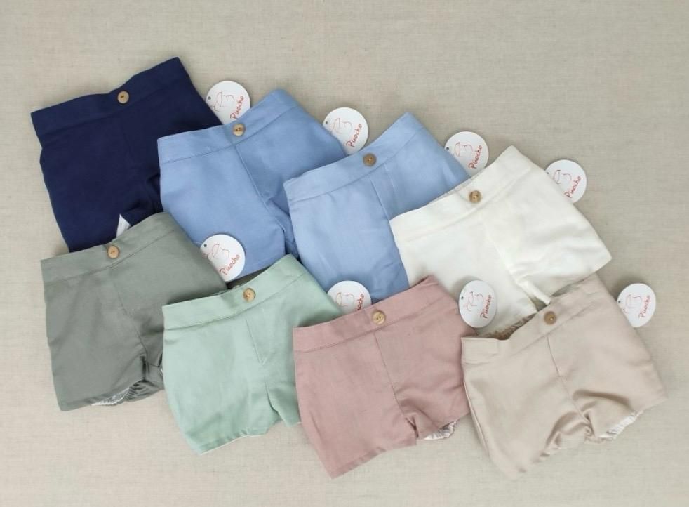 Nueve pares de pantalones cortos coloridos para bebé en varios tonos, cada uno con un botón y una etiqueta blanca.