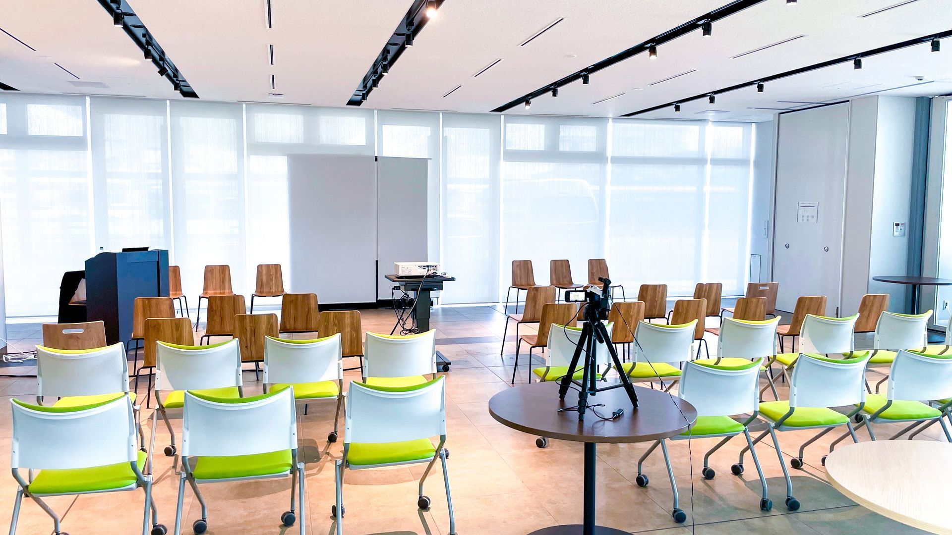 Salle de conférence moderne avec des rangées de chaises colorées et un trépied centré sur un parquet.