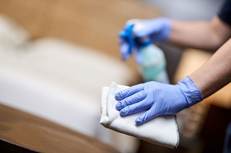 Des mains gantées de bleu essuient une surface en bois avec un chiffon blanc et un flacon pulvérisateur à proximité