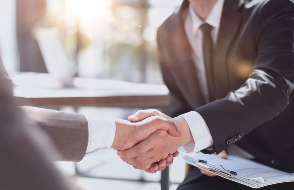 Dos hombres de traje se dan la mano sobre una mesa; uno de ellos sostiene un portapapeles.