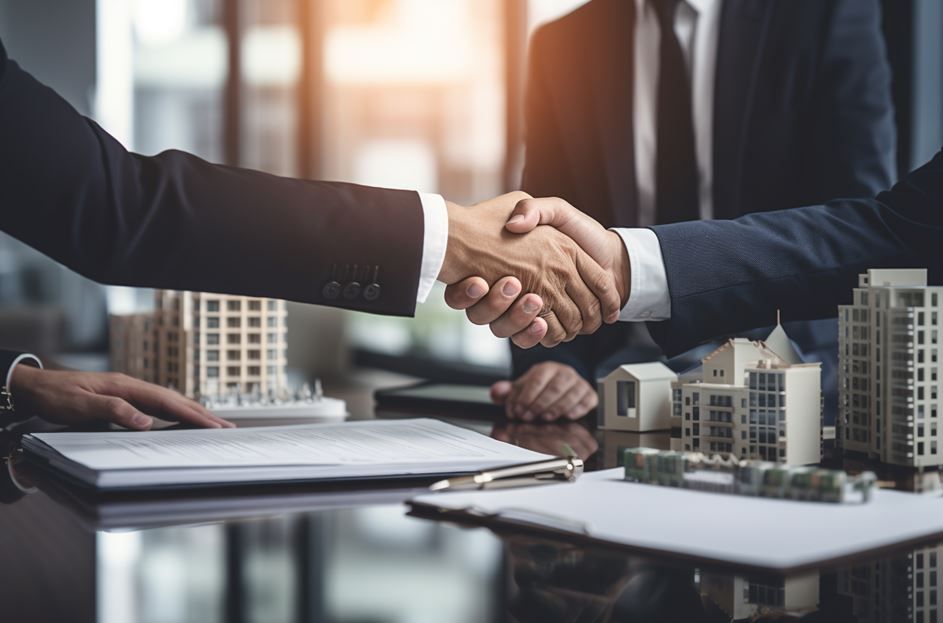 Dos hombres de negocios se dan la mano sobre una mesa con maquetas y documentos arquitectónicos.