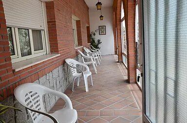 Una fila de sillas blancas están sentadas en un porche al lado de una pared de ladrillos.