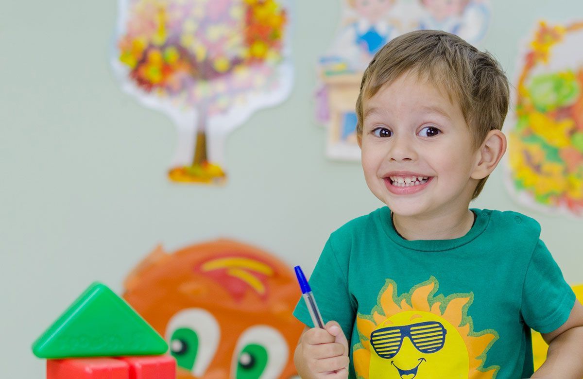 Ein kleiner Junge trägt ein grünes Shirt mit einer Sonne darauf, hält einen Stift und lächelt.