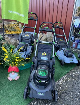 Trois tondeuses à gazon alignées sur l'herbe, le long d'une clôture rouge, avec un pot de fleurs et des éléments de décoration de jardin.
