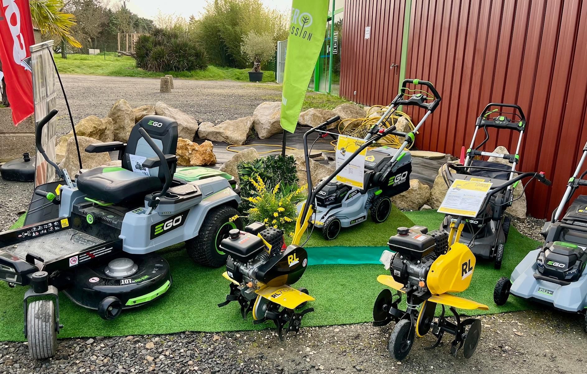 Assortiment de tondeuses à gazon et d'équipements de jardin exposés à l'extérieur sur du gravier, à côté d'une clôture rouge.