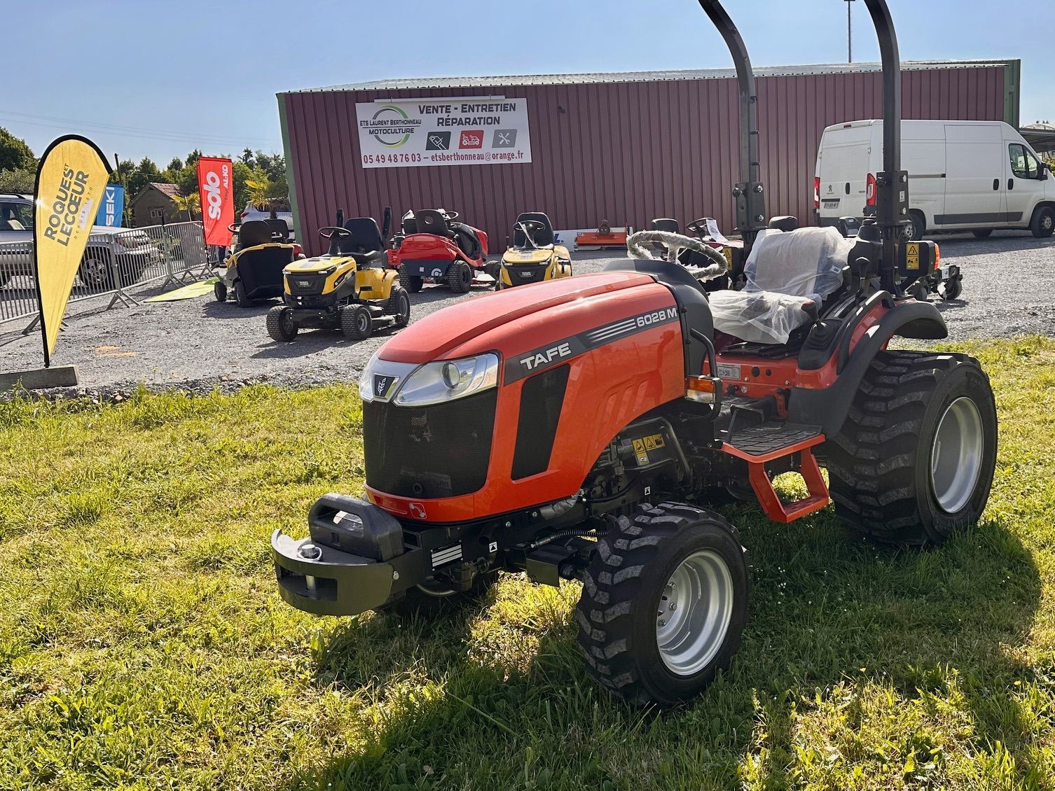 Tracteur tondeuse orange stationné sur l'herbe d'un terrain de matériel de jardinage, avec d'autres tondeuses et un bâtiment derrière.