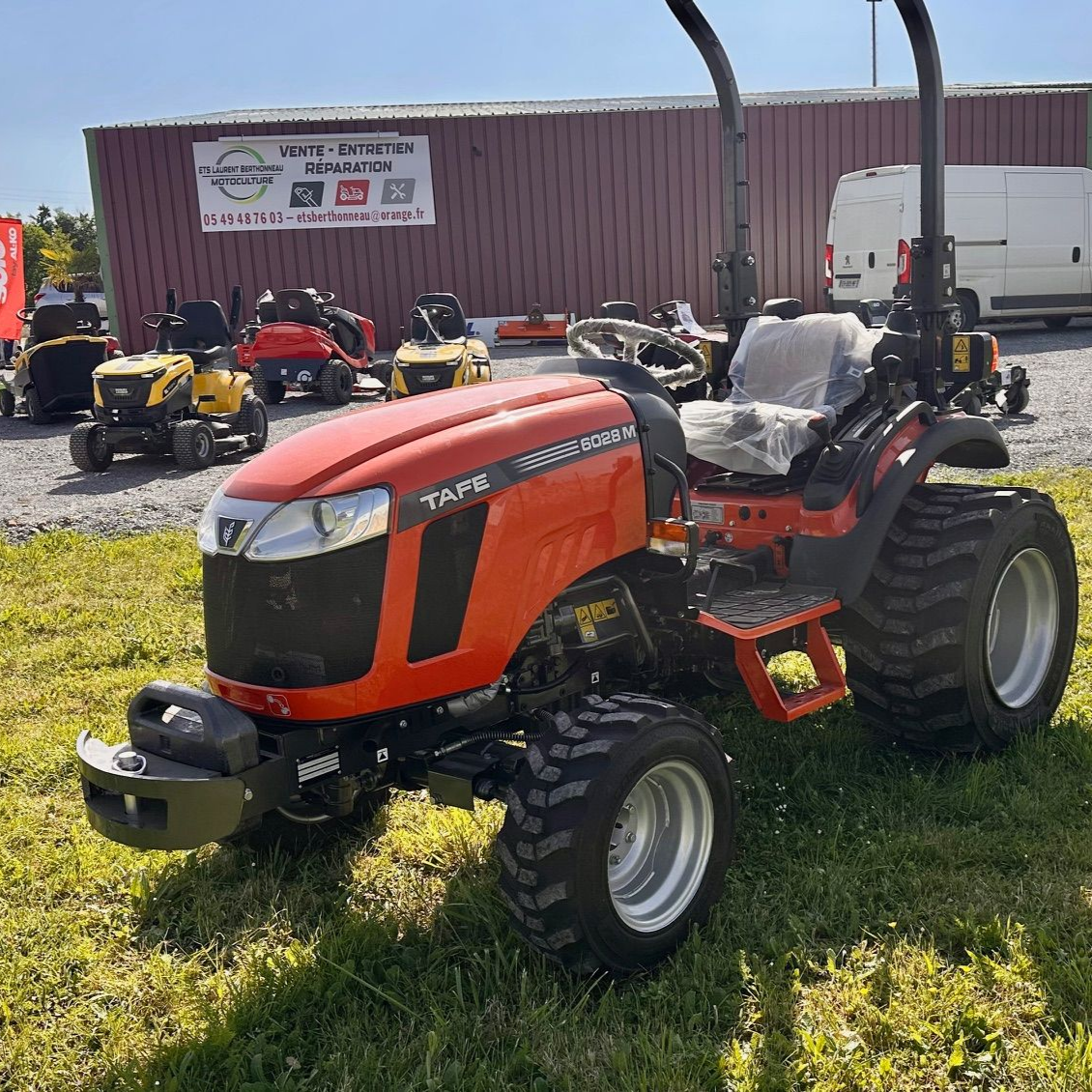 Tracteur orange stationné sur l'herbe d'un terrain de matériel de plein air, avec d'autres machines et un bâtiment derrière.