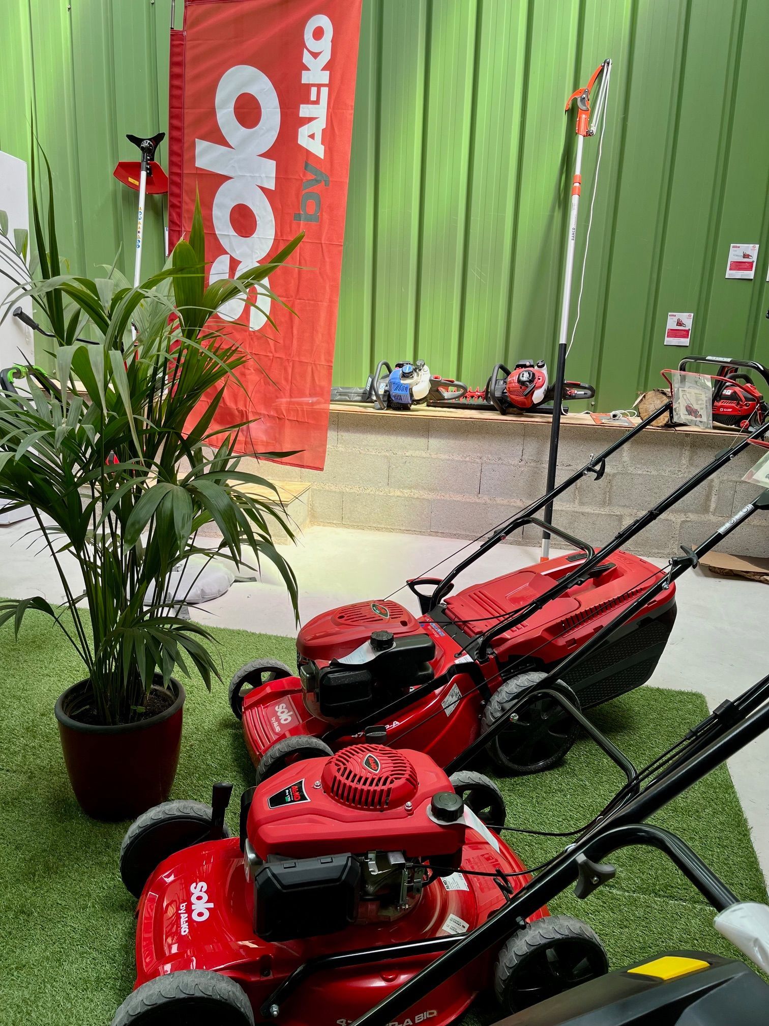 Tondeuses à gazon rouges sur gazon artificiel à côté d'une plante en pot et d'une banderole orange dans une salle d'exposition