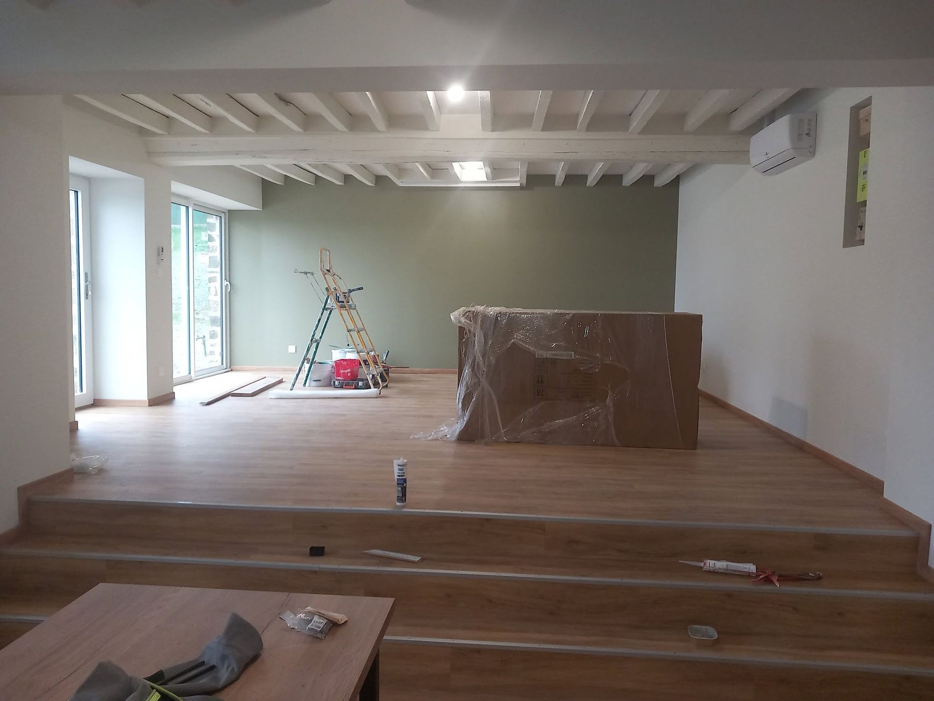 Une pièce en cours de rénovation avec un parquet, des poutres blanches au plafond et un mur d'accent vert sauge.