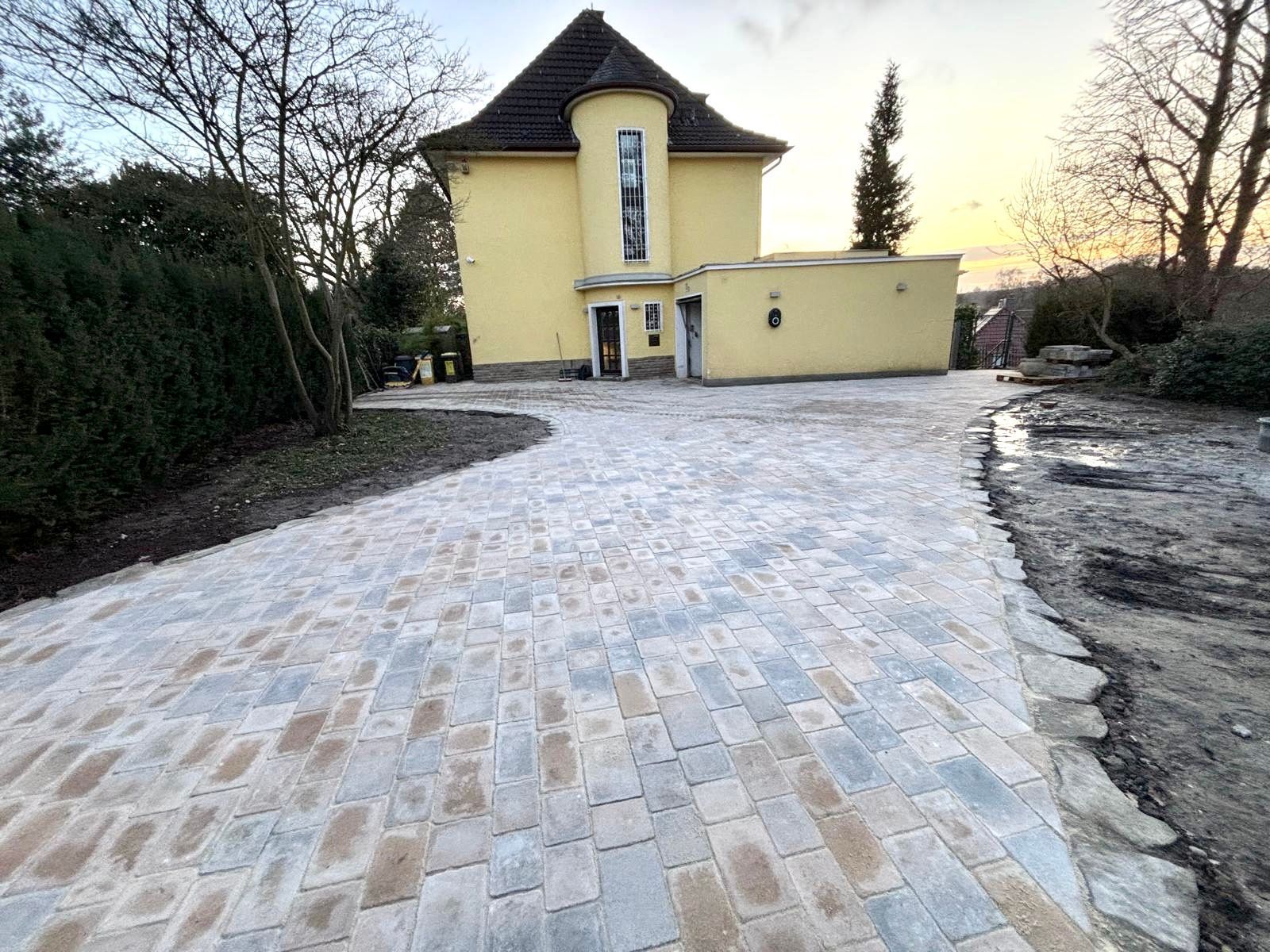 Gelbes Haus mit gemauerter Auffahrt, umgeben von einer gepflegten Gartenanlage, unter einem bewölkten Himmel