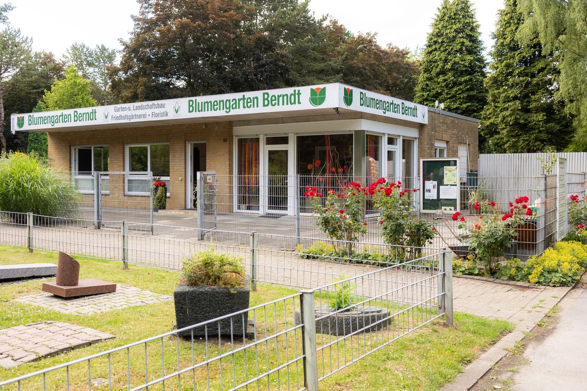 Außenansicht des Gebäudes „Blumengarten Bernd“ mit Beschilderung, roten Rosen und einem Zaun