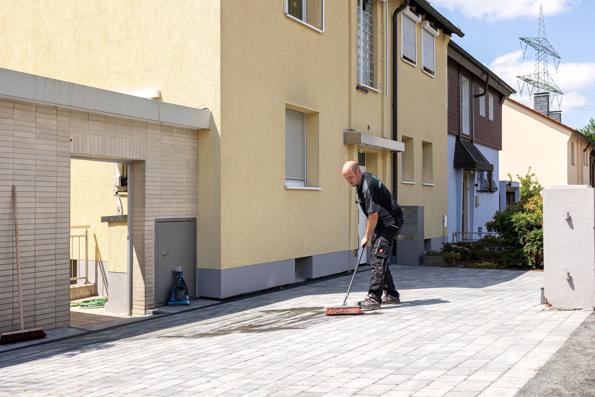 Ein Arbeiter fegt eine gepflasterte Auffahrt vor einem gelben Gebäude mit offener Garage