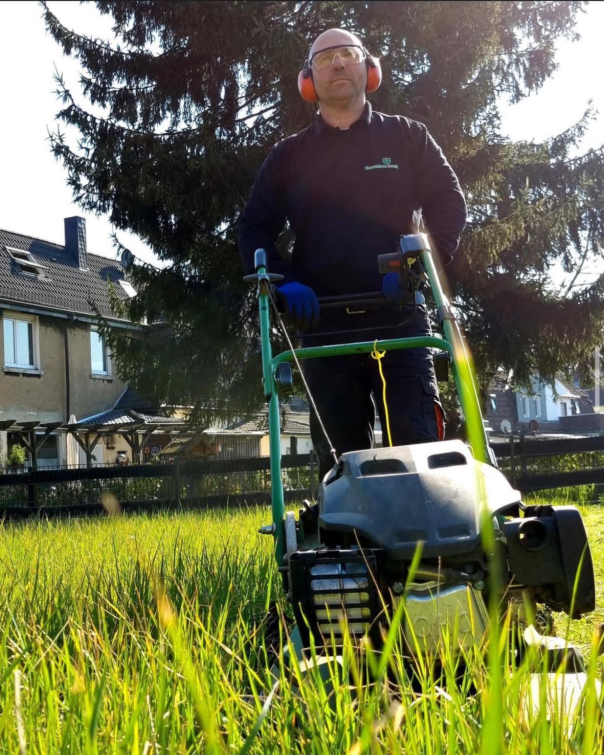 Ein Gärtner mit Gehörschutz und Handschuhen mäht mit einem grün-grauen Rasenmäher den Rasen vor einem Haus