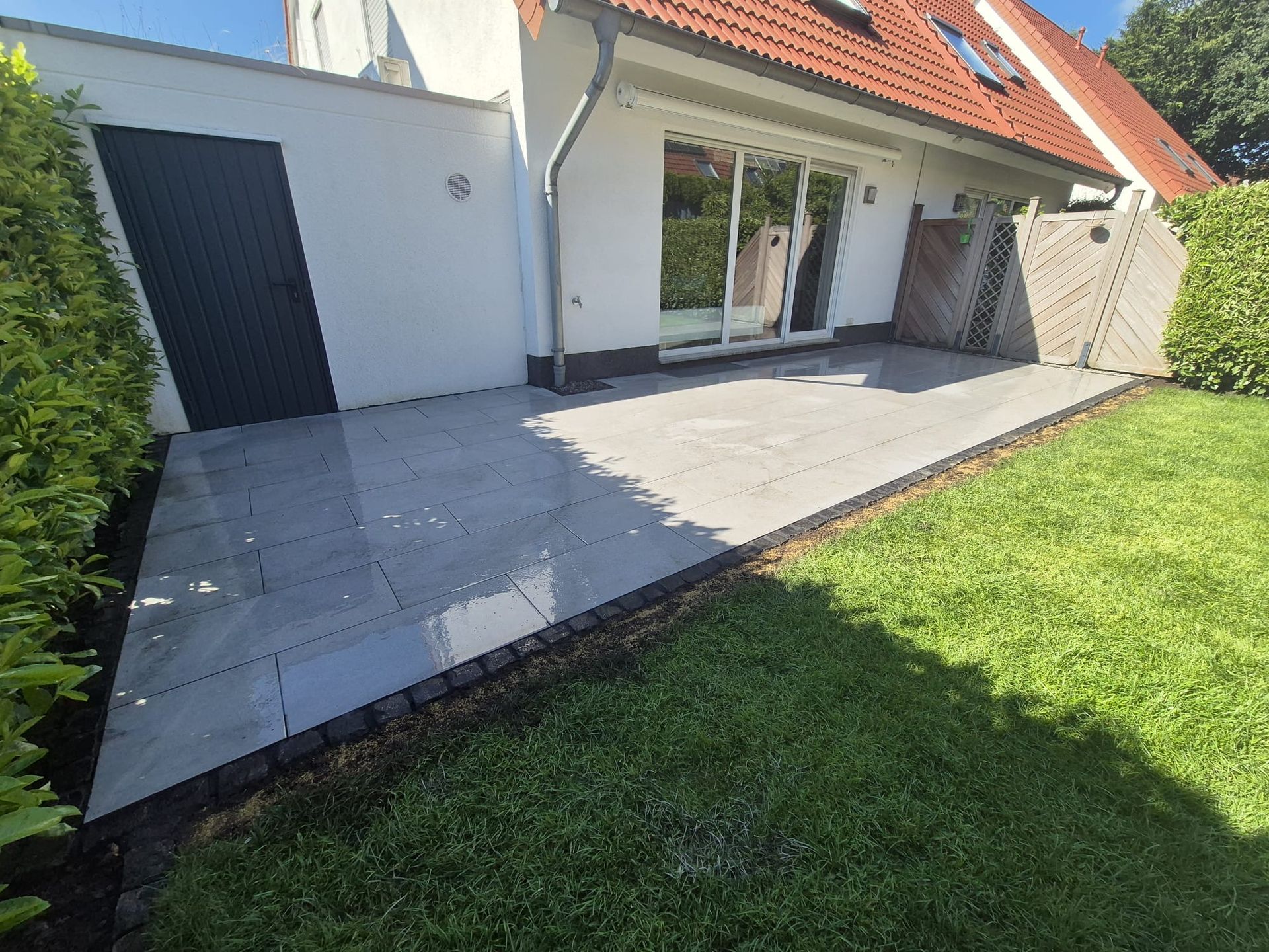 Terrasse mit grauen Fliesen, eingefasst von dunklem Stein, grünem Gras und einem weißen Haus mit Glasschiebetüren