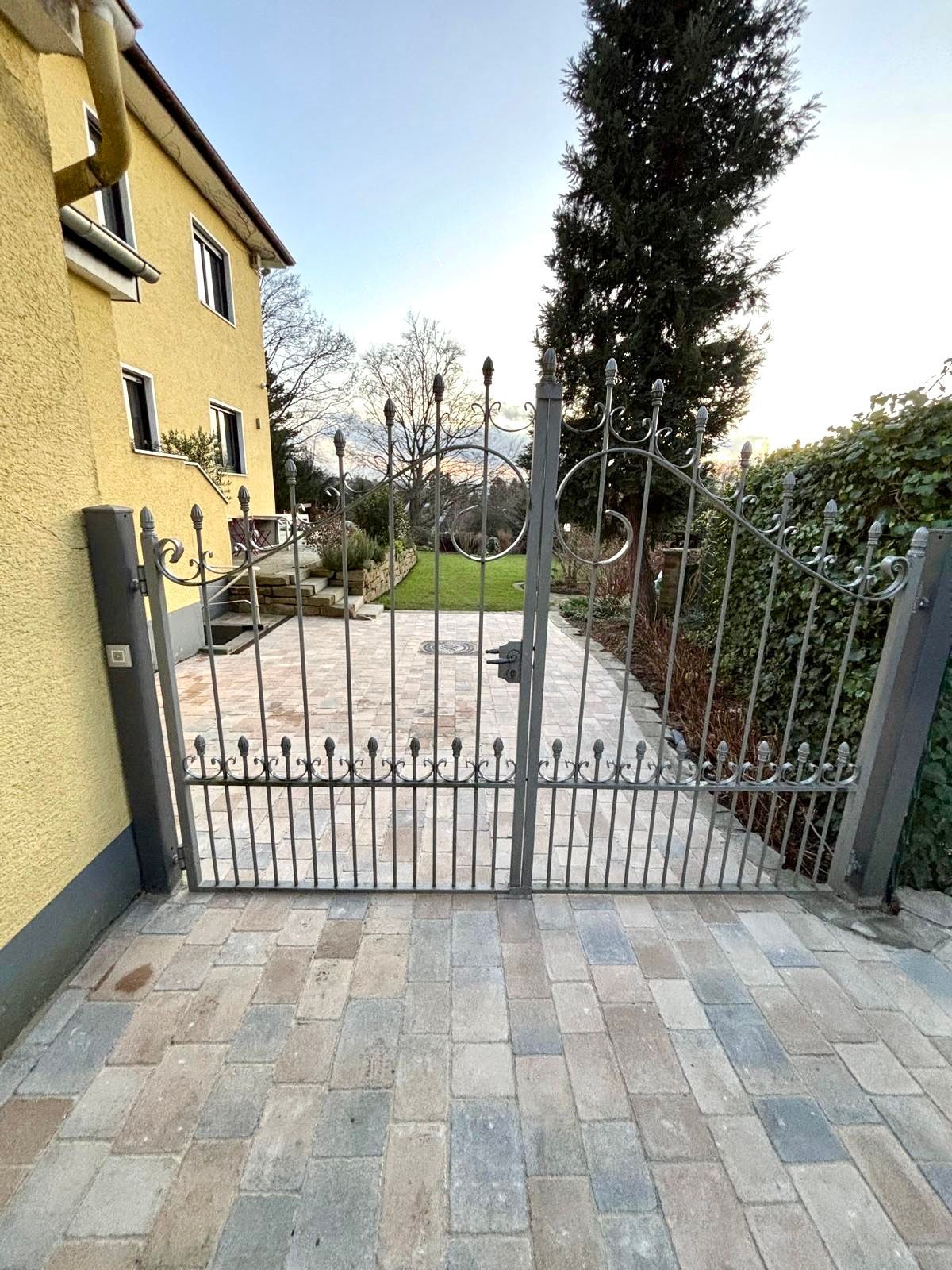 Ein offenes Metalltor mit Zierde steht auf einem Backsteinweg. Im Hintergrund sind ein gelbes Haus und Grünpflanzen zu sehen