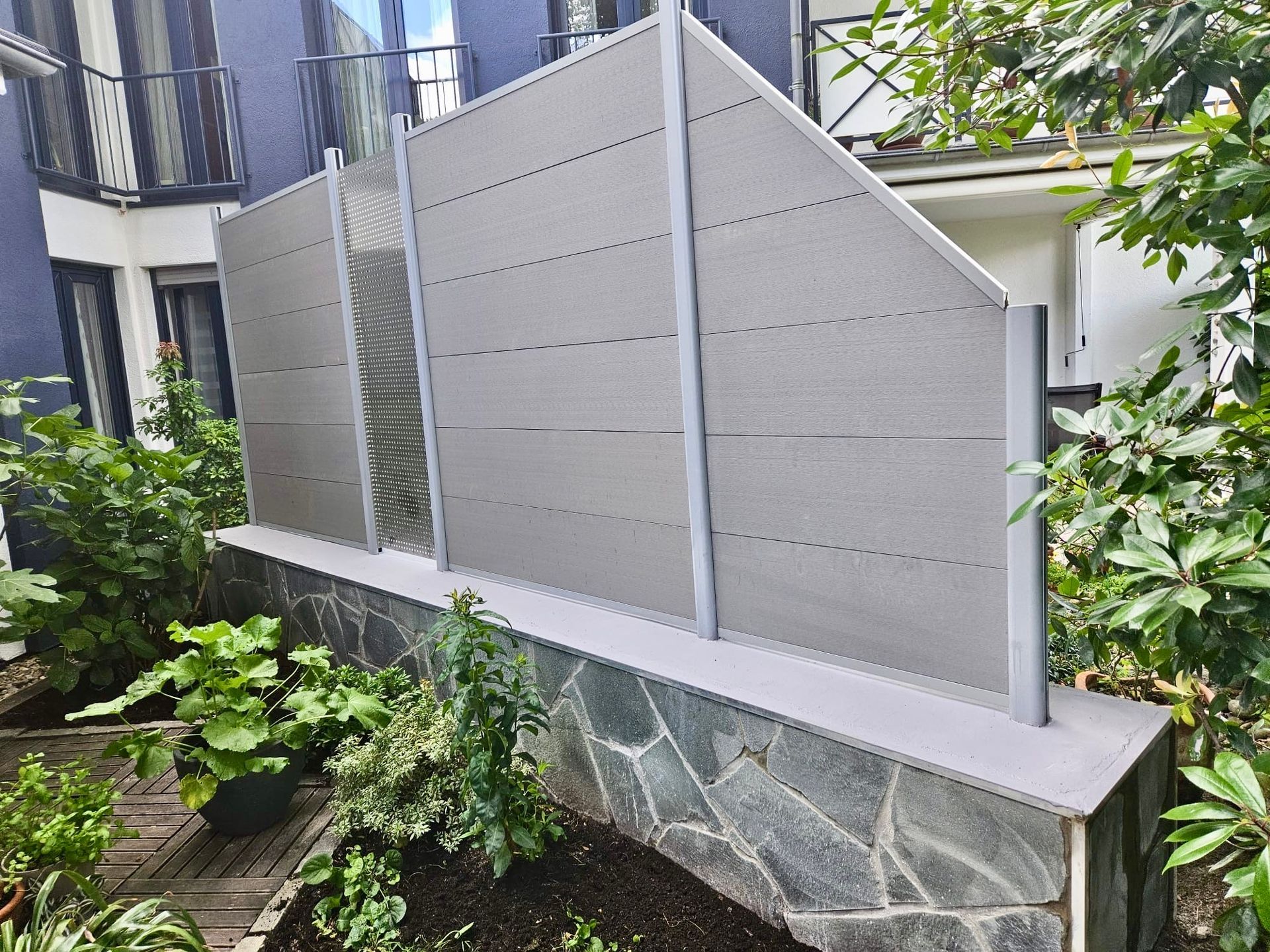 Grauer Sichtschutzzaun auf einer Steinmauer, umgeben von Grün