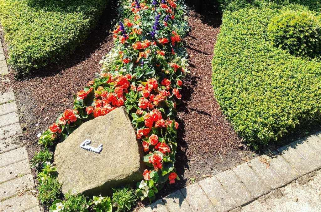 Grab mit einem Steinmarker, orange und blaue Blumen bedecken das Grab und mit Büschen und bildet es ein Muster