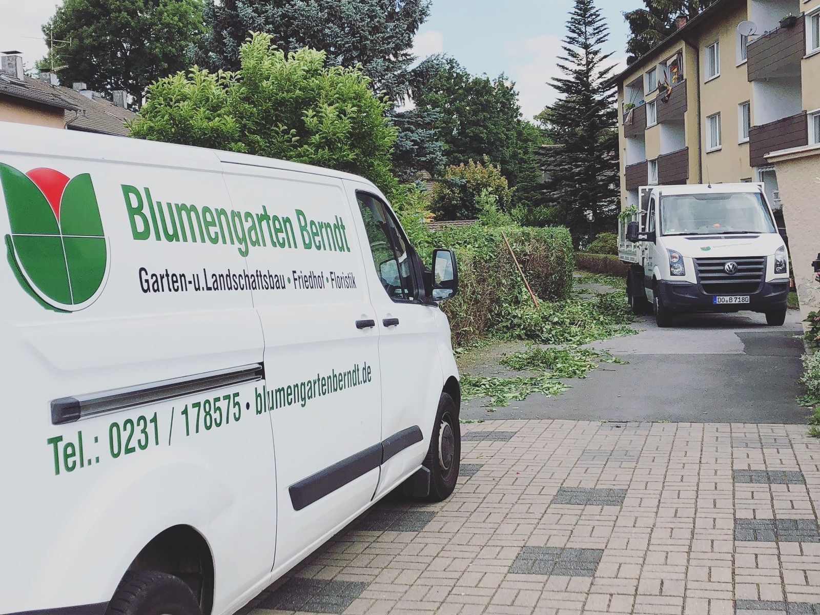 Zwei weiße Lieferwagen von „Blumengarten Berndt“ stehen vor einer Wohnsiedlung und kümmern sich um den Grünschnitt