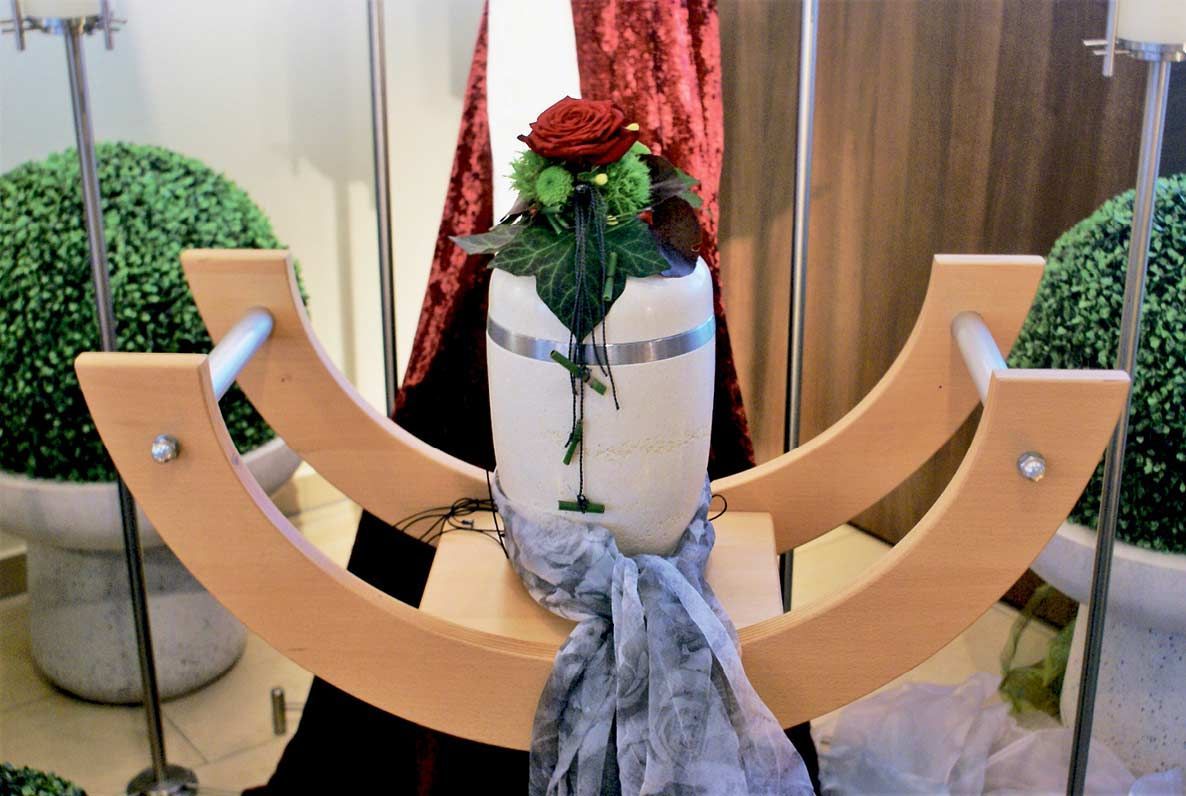 Eine weiße Urne mit einer roten Rose darin steht auf einem Holztisch.