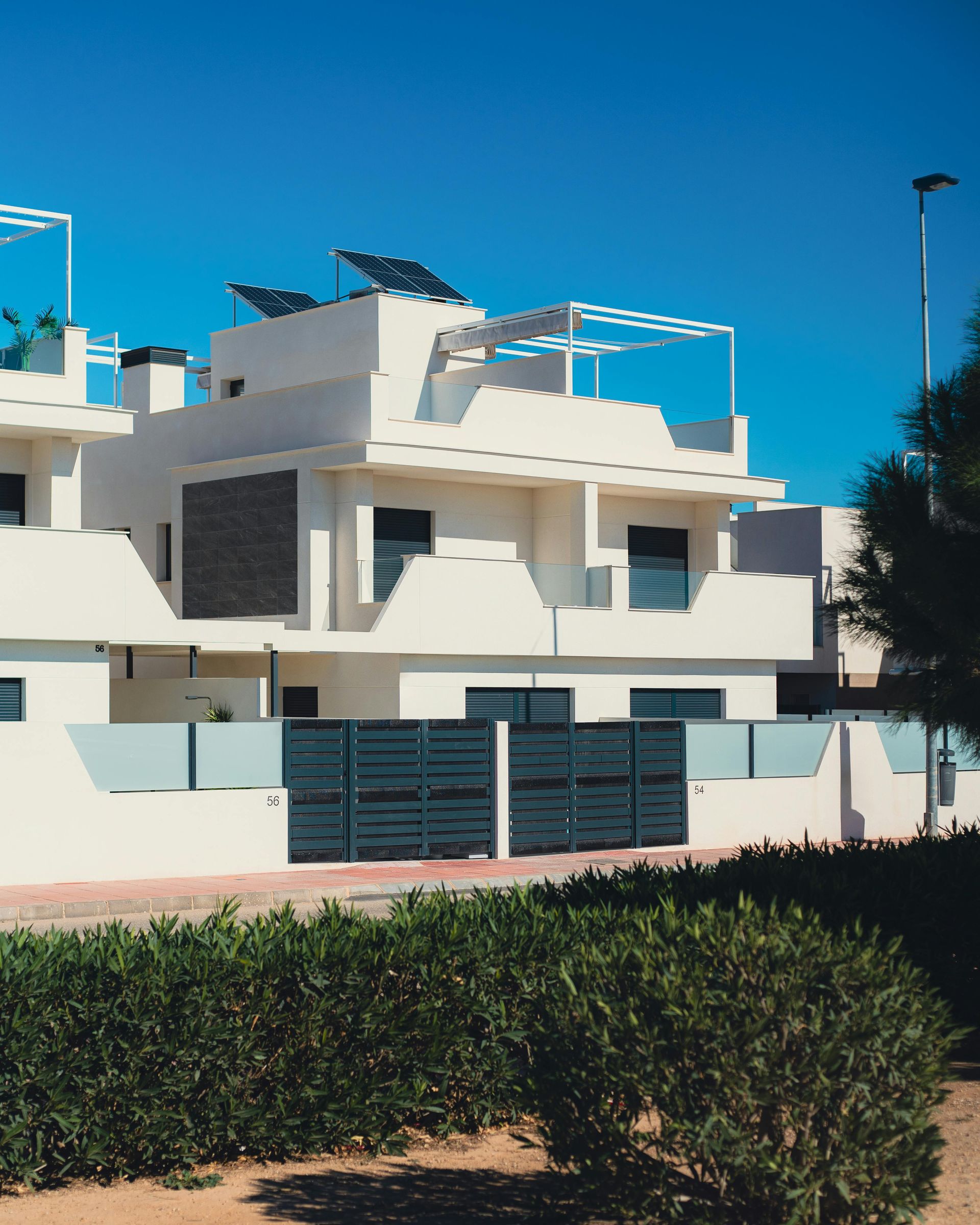 Casa moderna blanca con paneles solares en el tejado detrás de un seto bajo un cielo azul despejado.