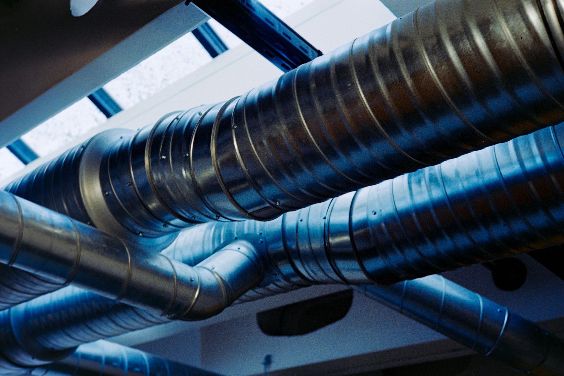 Conductos de ventilación metálicos que atraviesan un techo en un interior industrial, iluminados con luz azul fría.