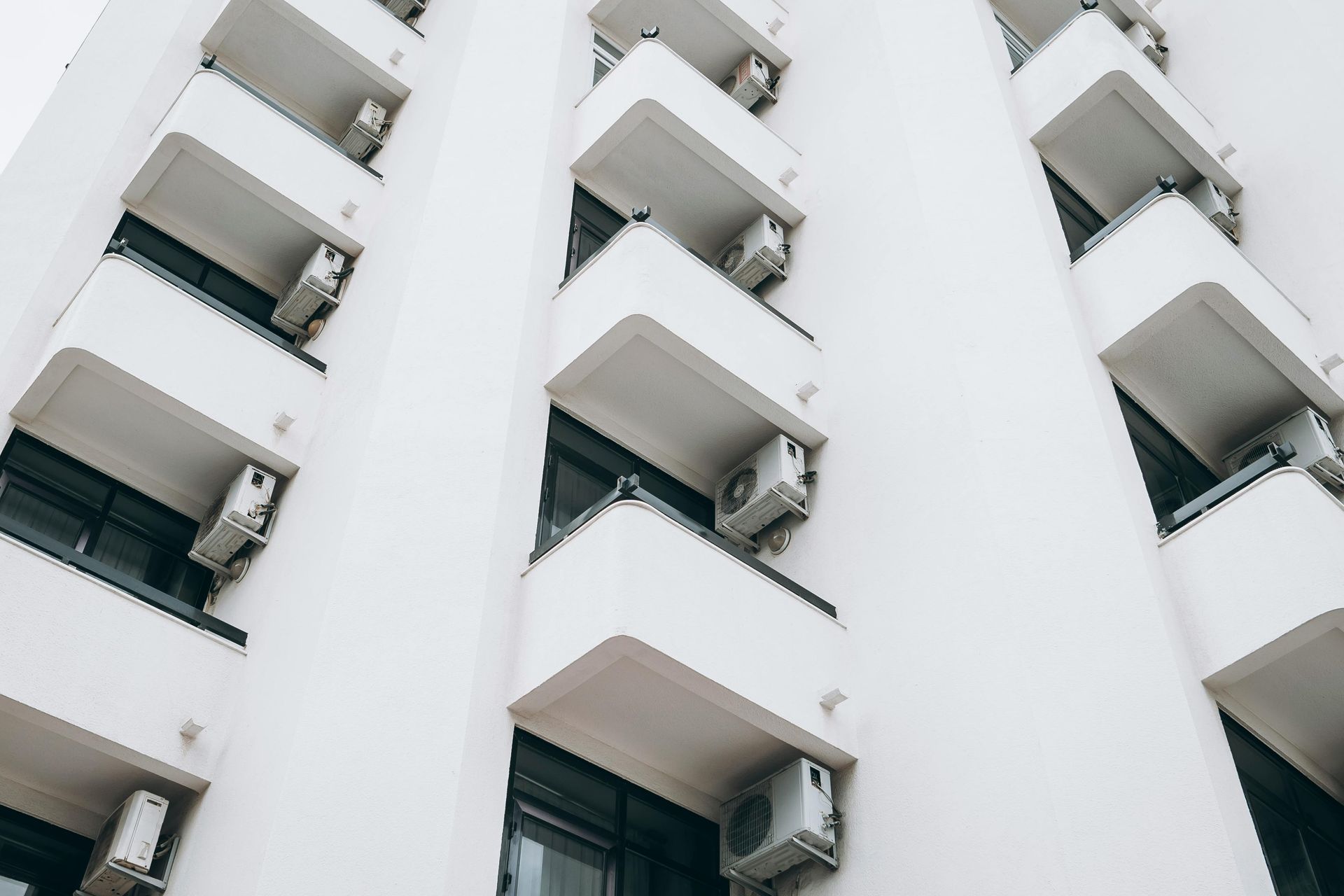 Fachada blanca de un edificio de apartamentos con hileras de balcones salientes y ventanas oscuras.