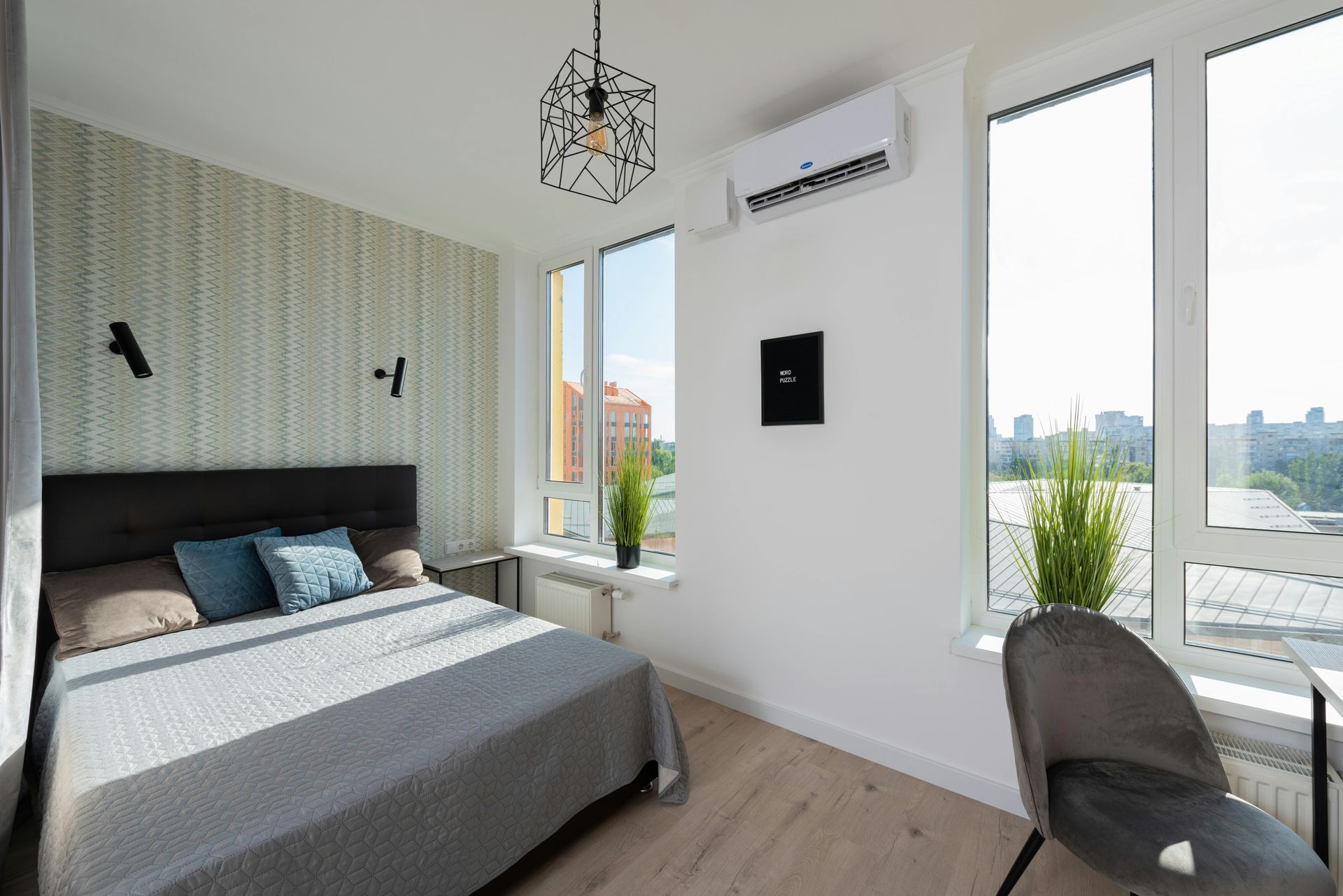 Habitación luminosa con cama gris, cabecero estampado, grandes ventanales y vistas a la ciudad.