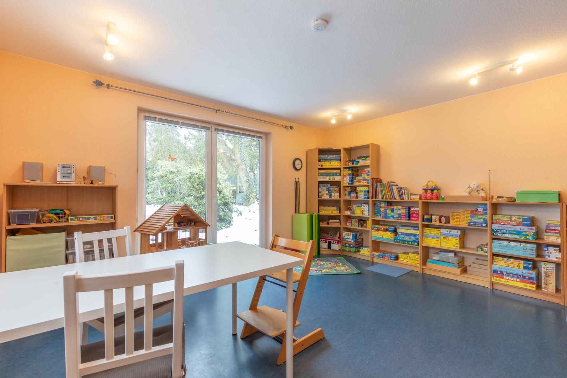Helles Klassenzimmer mit weißen Tischen, Holzstühlen, Bücherregalen und einer großen Glastür zum Garten.