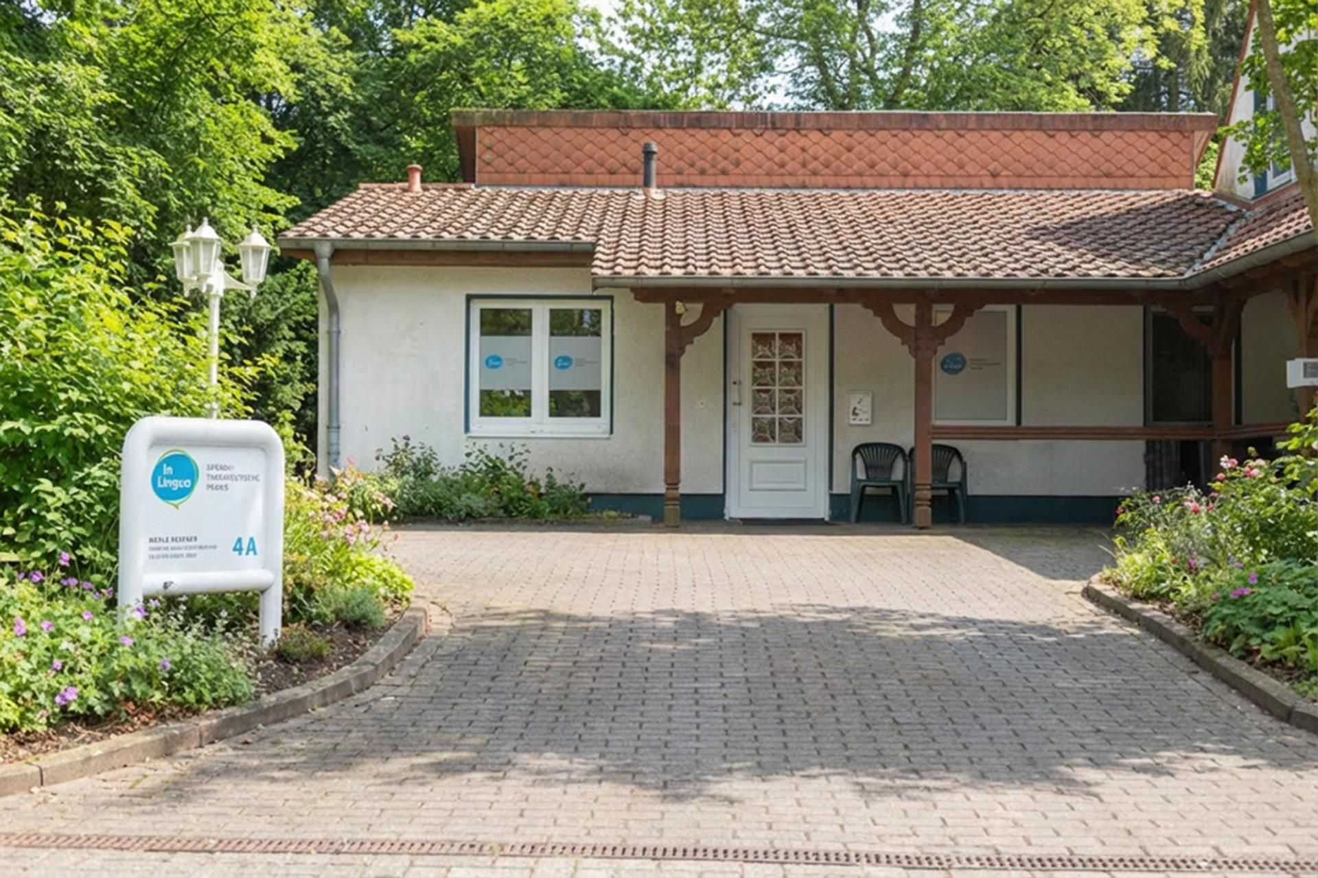 Kleiner Gebäudeeingang mit Ziegeldach, weißem Schild und gepflasterter Auffahrt, umgeben von Grün.