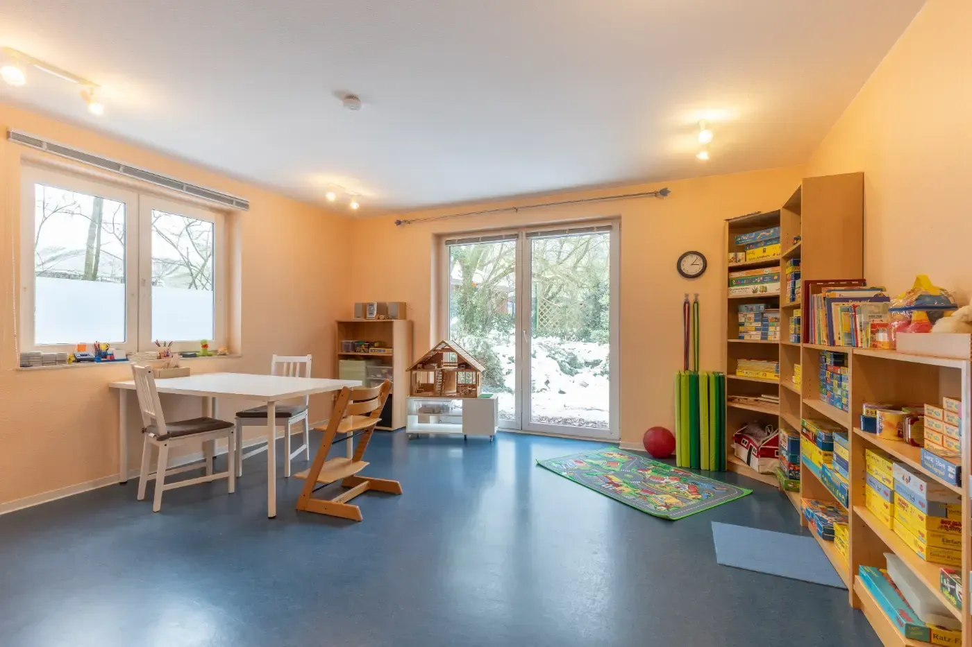 Helles Spielzimmer mit blauem Boden, Spielzeugregalen, Tisch und Glasschiebetür zum Garten