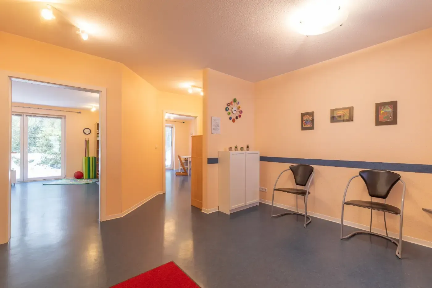 Heller Flur mit pfirsichfarbenen Wänden, grauem Boden, Stühlen auf der rechten Seite und Tageslicht aus einem Zimmer mit Fenster.