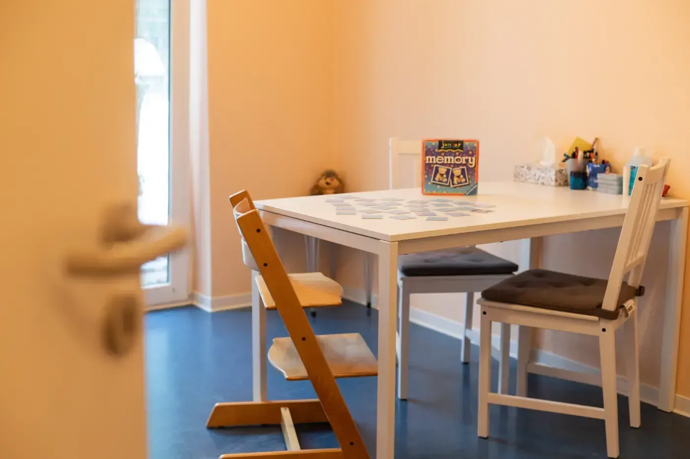 Kleiner Essbereich mit einem weißen Tisch, Stühlen unterschiedlicher Größe und einem Kochbuch darauf, durch einen Türspalt zu sehen.