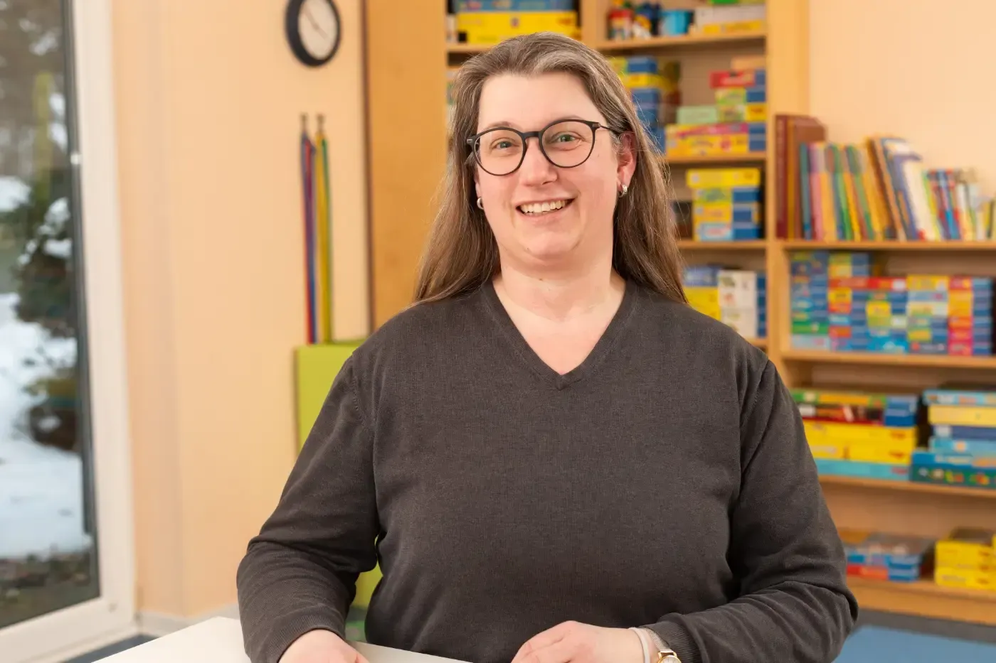 Eine lächelnde Person mit Brille steht in einem farbenfrohen Klassenzimmer mit Regalen voller Bücher und Lernmaterialien.