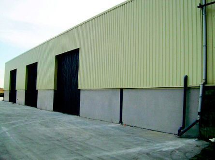 Exterior de una nave industrial con revestimiento verde pálido, puertas negras y una zona de carga de hormigón.