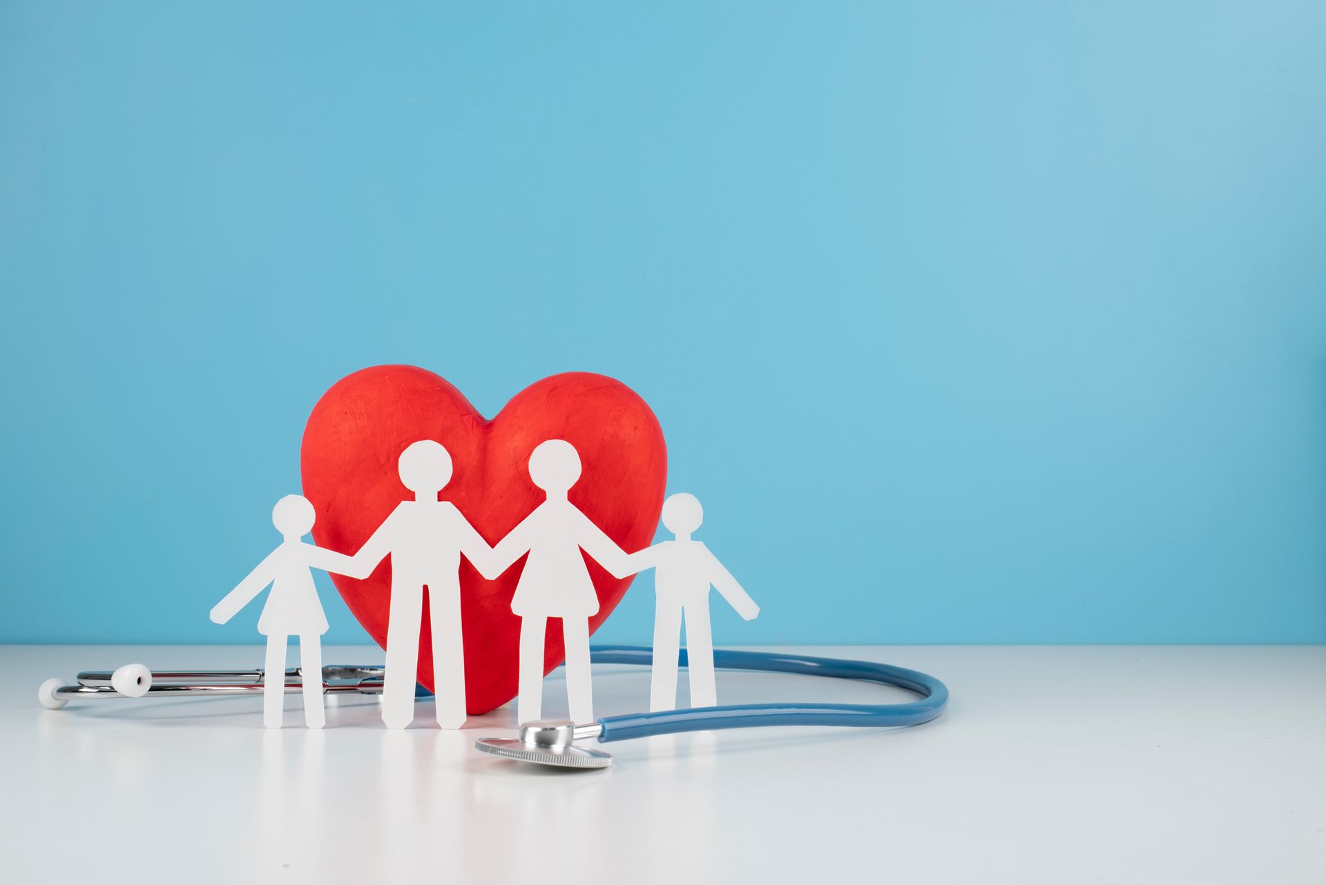Figuras recortadas de una familia tomadas de la mano delante de un corazón rojo y un estetoscopio sobre un fondo azul.