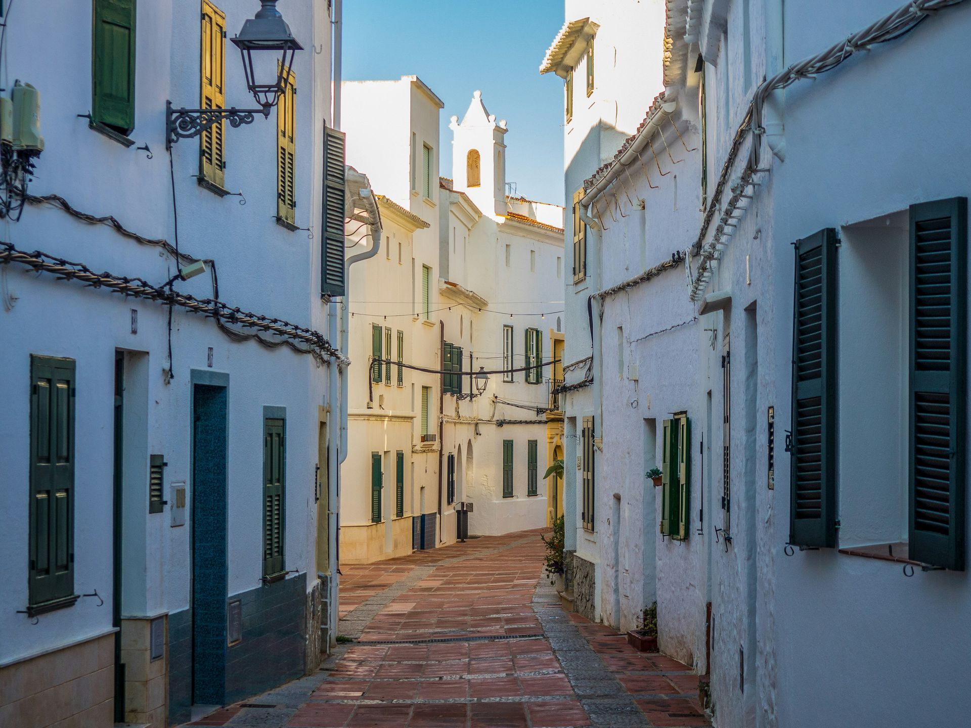 Calle estrecha adoquinada con edificios blancos, contraventanas verdes y una torre de iglesia en la distancia.