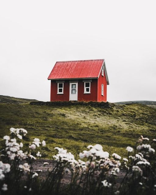 Casita roja con ventanas blancas en una colina cubierta de hierba bajo un cielo nublado, con flores blancas delante.