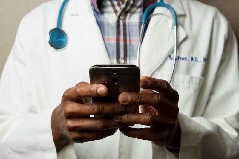 Médico con bata blanca sosteniendo un teléfono inteligente, con un estetoscopio alrededor del cuello.