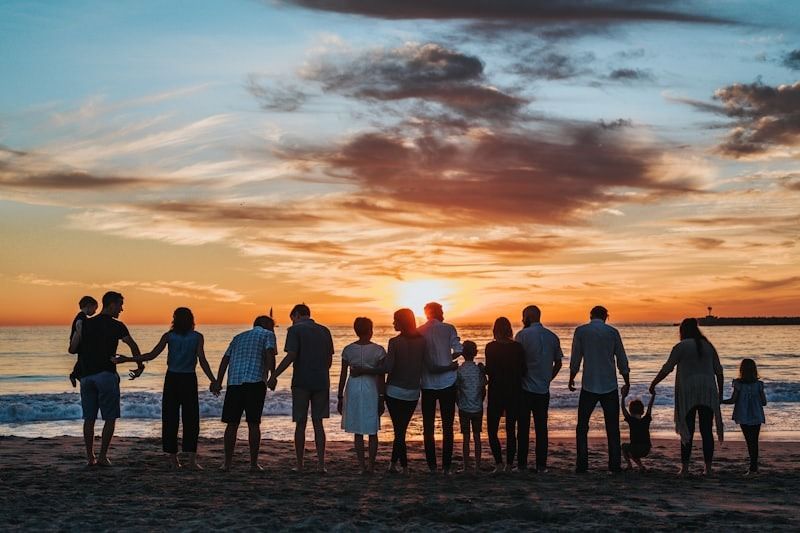 Personas tomadas de la mano en una playa al atardecer, frente al océano bajo un cielo colorido.