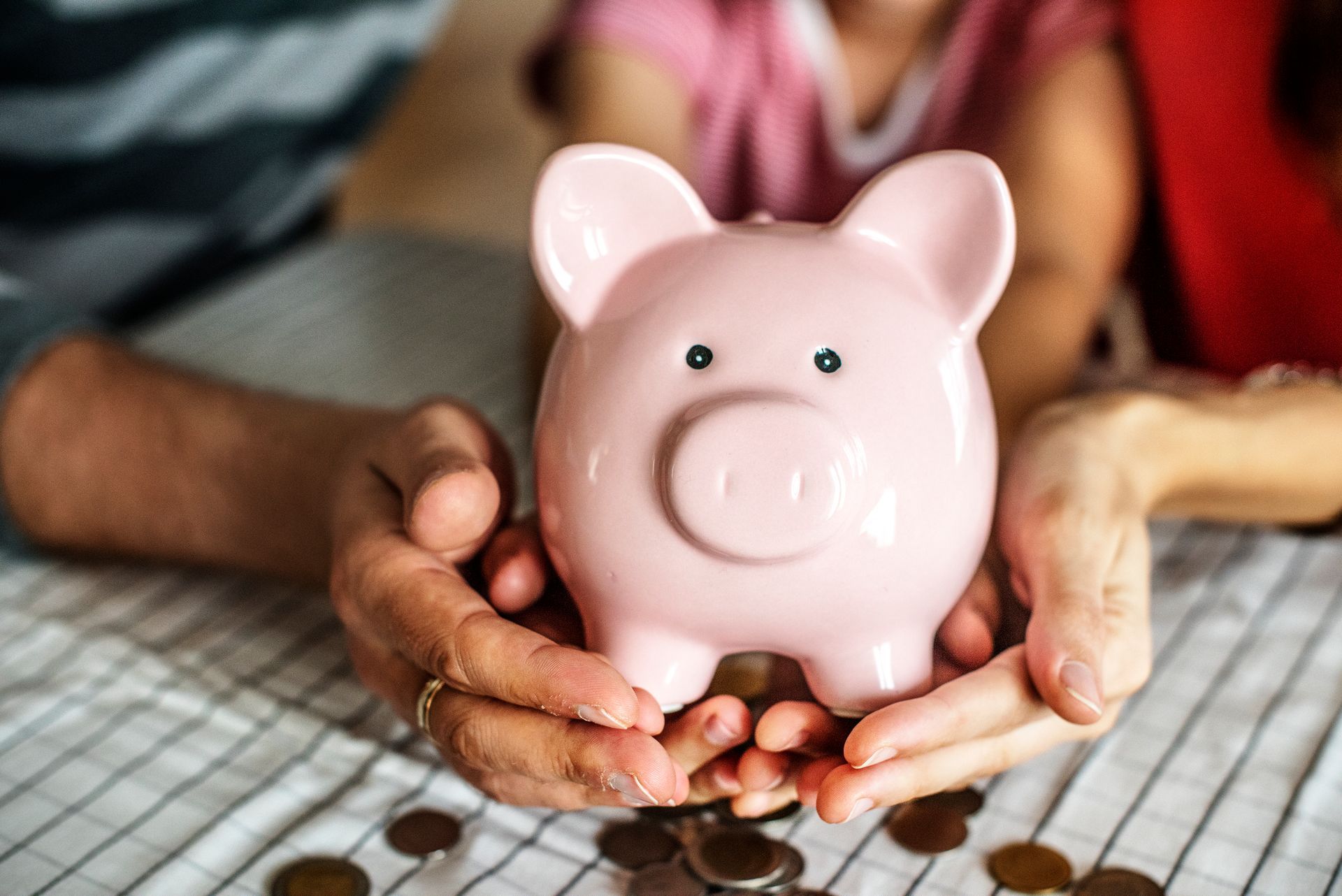 Manos sosteniendo una alcancía rosa; monedas esparcidas sobre una superficie a cuadros.