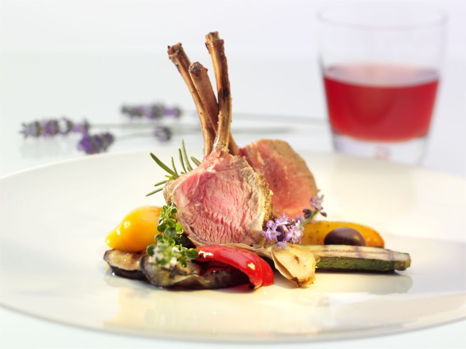 Lammkotelett mit geröstetem Gemüse, garniert mit Kräutern, auf einem weissen Teller, im Hintergrund ein Getränk.