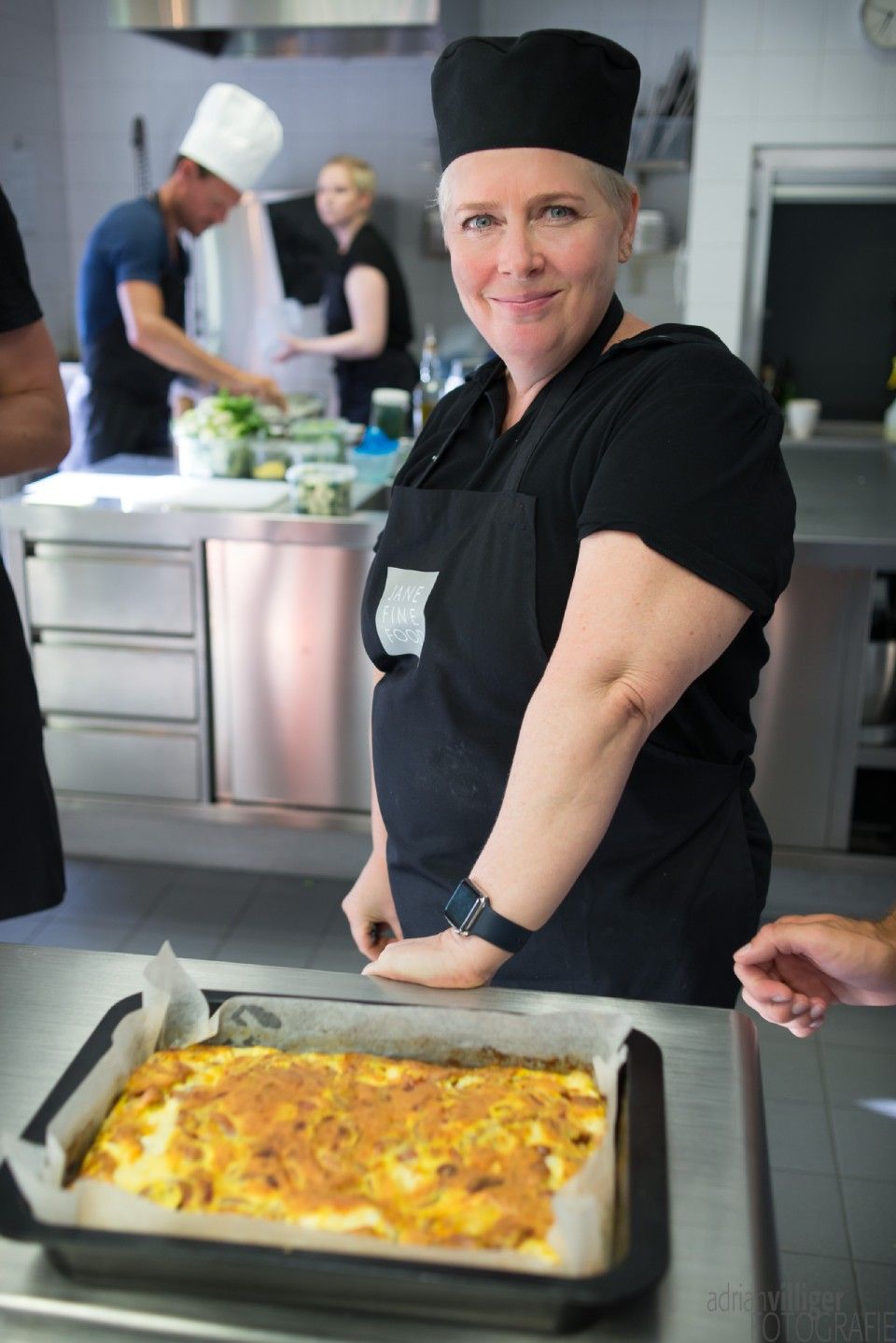 Frau mit Kochmütze und Schürze in der Küche, lächelnd, ein Teller vor sich. Weitere Köche im Hintergrund.