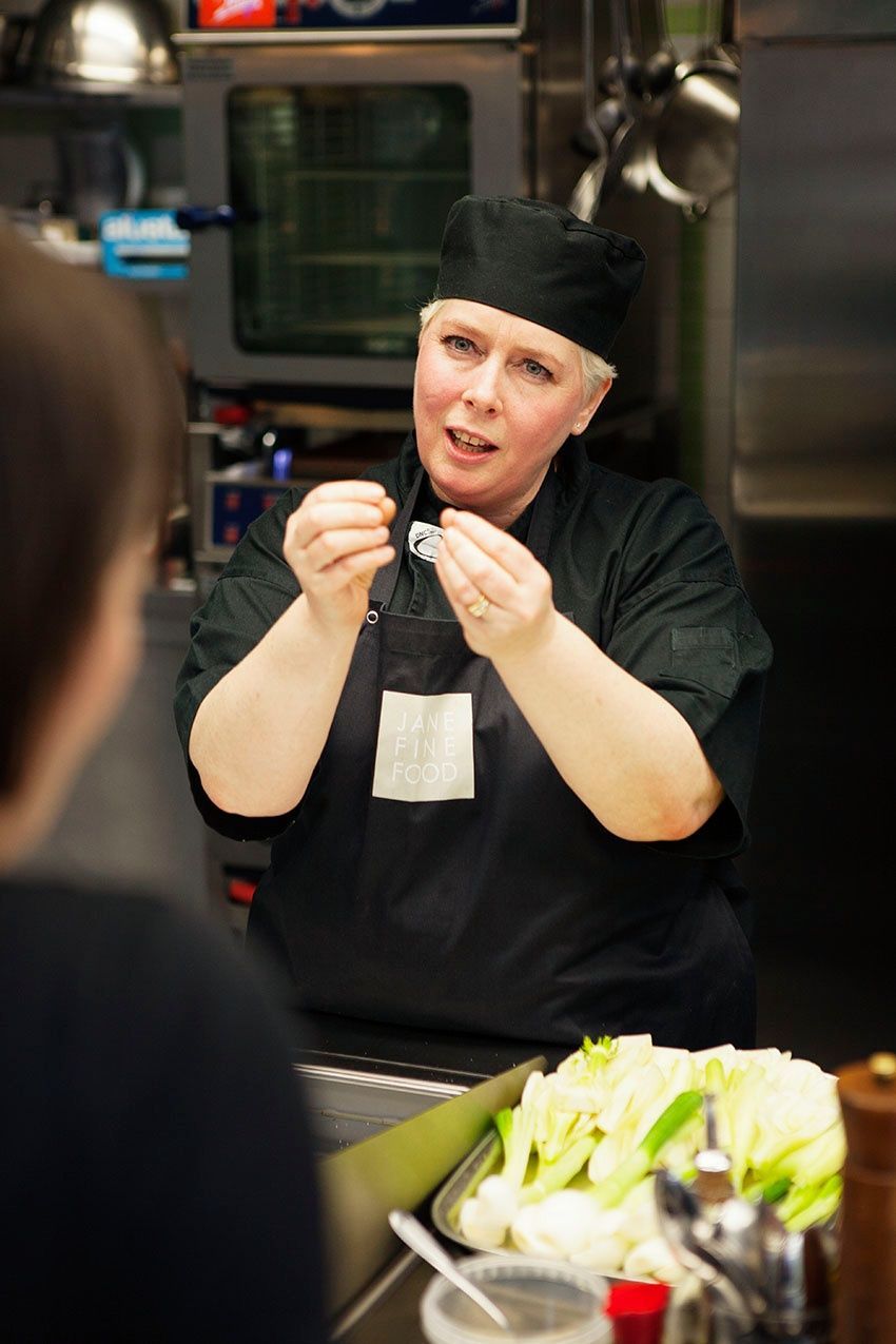 Ein Koch in schwarzer Uniform erklärt einer Person in einer Grossküche etwas; er hält dabei kleine Gegenstände in der Hand.