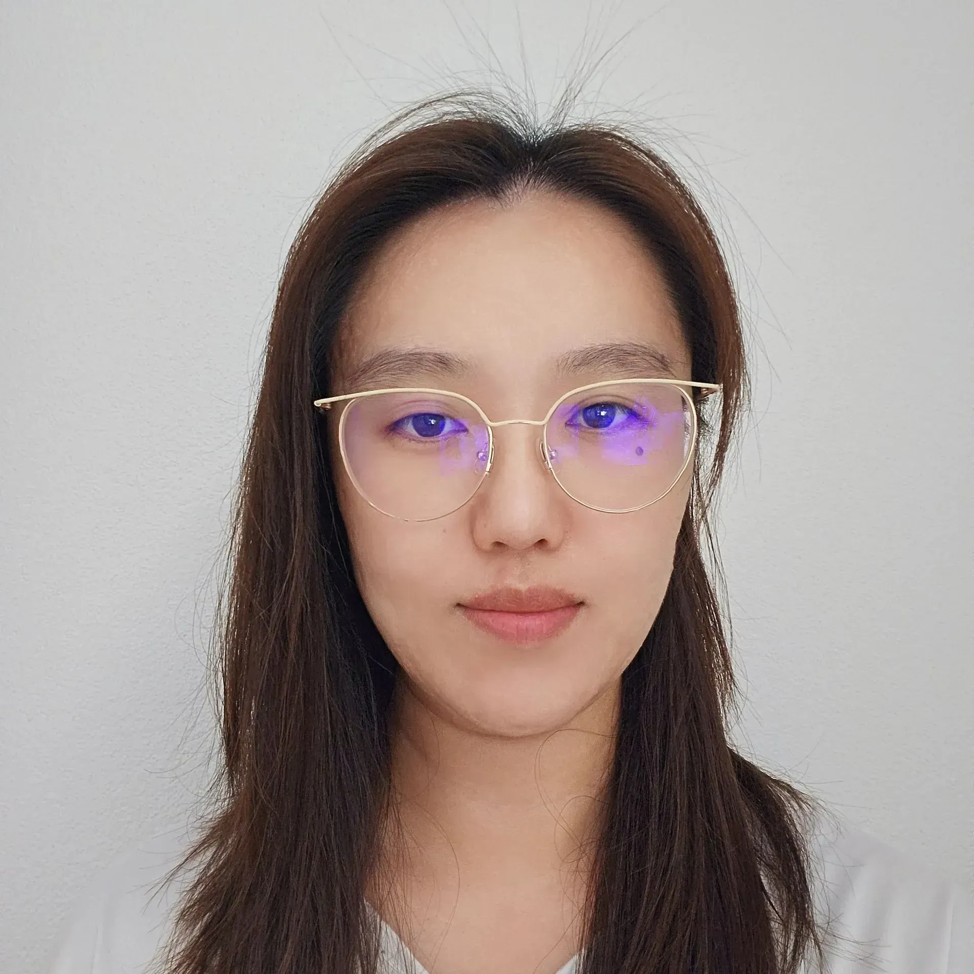 Frau mit Brille schaut in die Kamera. Langes dunkles Haar, neutraler Gesichtsausdruck, Inneneinrichtung.