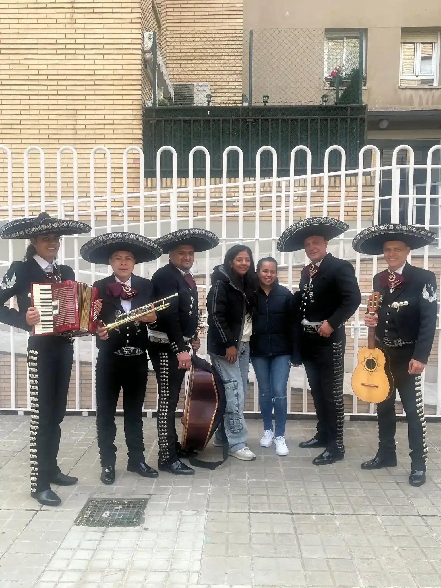 Banda de mariachis con dos mujeres frente a un edificio con una valla.