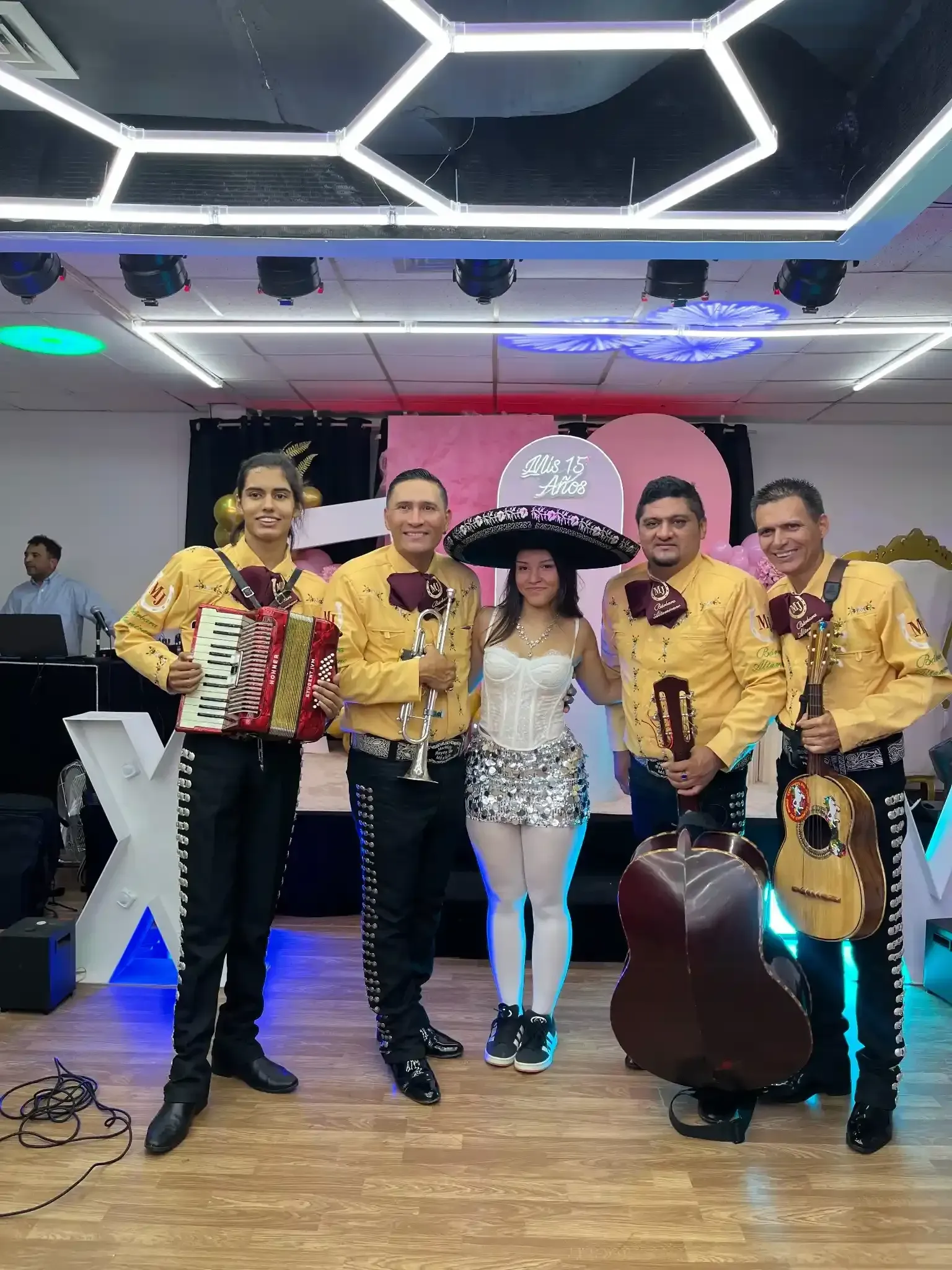 Banda de mariachis con acordeón, trompetas, guitarra y cantante en un lugar decorado.