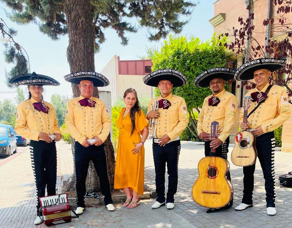 Banda de mariachis con una mujer vestida de amarillo posando afuera cerca de árboles y un edificio.